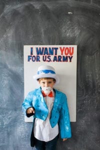 Child dressed as Uncle Sam in front of an I Want You for U.S. Army sign