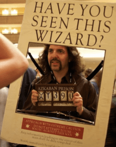 Person framed by a Have You Seen This Wizard poster, holding a prisoner number sign